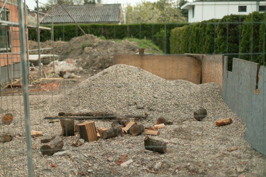 Foreground gravel and chopped logs with large gravel pile nearby, temporary wooden formwork, and scaffolding visible within fenced residential construction area.