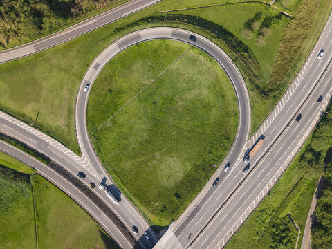 Aerial view of a highway interchange with a circular loop ramp surrounded by green fields and trees in Brazil.