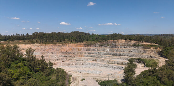 Aerial view of an open-pit quarry with terraced rock faces and exposed stone, surrounded by dense green forest under a clear blue sky in Brazil.