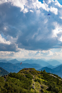 Aerial view of paraglider above the mountain landscape of Konigssee, with a windsock on a grassy peak and distant blue mountains in Schonau am Konigssee, Bavaria, Germany.