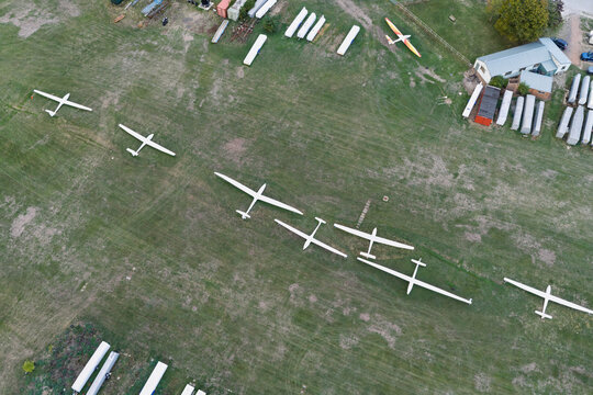 Aerial view of several white gliders parked in a row on a grassy airfield with hangars and buildings in the background in the United Kingdom.
