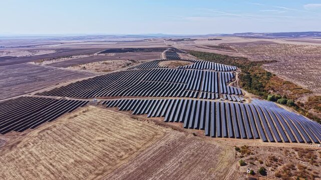 Rows of solar panels extend across farmland in a rural area, viewed from above during daylight, with fields and low hills around the installation.