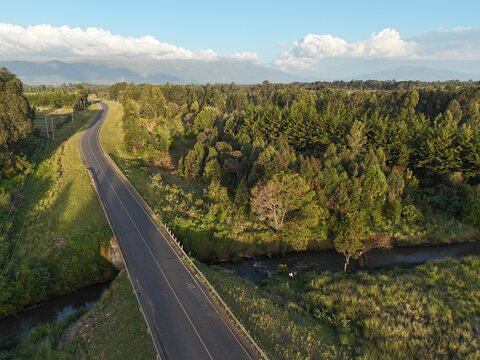 Aerial view of a winding asphalt road crossing a small stream, surrounded by dense green forests and rolling hills in Ol Kalou, Nyandarua County, Kenya.
