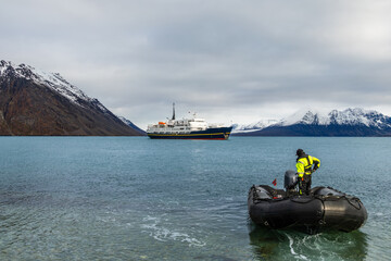 Person in an inflatable expedition zodiac boat approaching a passenger ship on calm Arctic water beneath snow-covered mountains, Krossfjorden, Spitsbergen, Svalbard, Norway © vadim.nefedov
