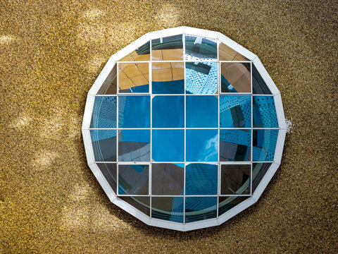 Aerial view of a circular glass skylight with a grid pattern set into a textured gravel surface, reflecting surrounding buildings in Belgrade, Serbia.