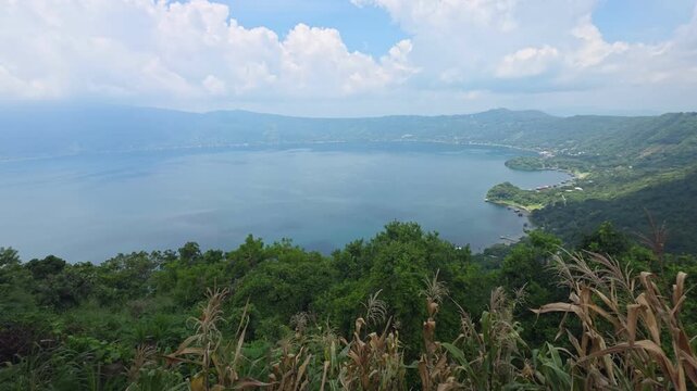 High-angle panoramic view of Coatepeque volcanic crater lake surrounded by lush green mountains and crops in El Salvador. Scenic volcanic landscape under a bright blue sky with white clouds, perfect f