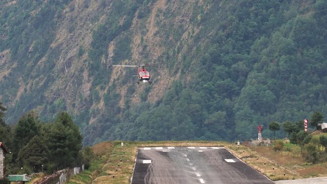 Red rescue helicopter approaching and landing on remote airstrip nestled in mountain valley, performing crucial maneuver in isolated and dangerous high altitude. Tengchong and Hillary Airport in Nepal