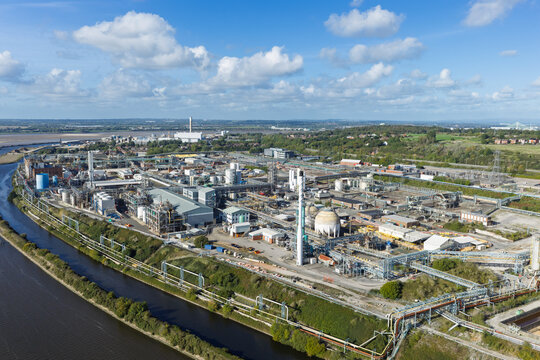 Aerial view of a large chemical industrial plant with storage tanks, complex piping, and processing facilities along a riverbank under a bright blue sky in the United Kingdom.