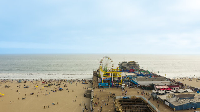 Aerial view of Santa Monica Pier with its Ferris wheel, roller coaster, and bustling boardwalk filled with people, extending into the Pacific Ocean in Santa Monica, California, United States.