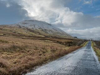 Snow-dusted hill looms over narrow road. Grassy fields border wet asphalt path. Sky cloudy with patches of blue. Scene feels quiet and remote. Ideal for travel, nature, or winter themes © mark_gusev
