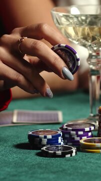 Hand placing poker chip beside stacked casino chips and playing cards on green felt with cocktail glass in background, close up gambling scene