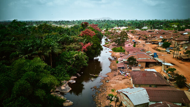 Aerial view of a river flowing through a village with lush tropical vegetation on one side and residential houses with metal roofs on the other in Idanre, Ondo, Nigeria.