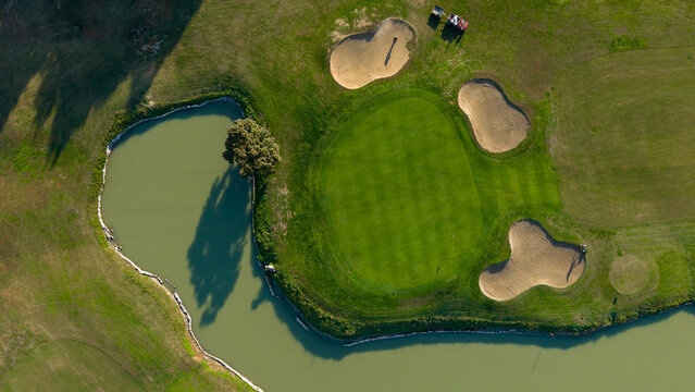 Aerial view of a golf course featuring a green putting area, sand bunkers, and a winding water hazard with a tree in Milan, Lombardy, Italy.