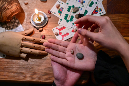 Person holding small beans and stones in hand during a traditional divination setup with playing cards and a lit candle. Fortune teller with cartomancy tools. Close-up of traditional mystical ritual