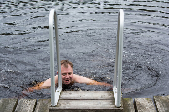 Man swimming in dark rippled water and approaching a wooden dock with a metal ladder