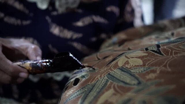 Close-up cinematic footage of artisan hands drawing traditional Indonesian batik pattern using hot wax canting tool on fabric. Authentic cultural heritage craftsmanship process in detail.