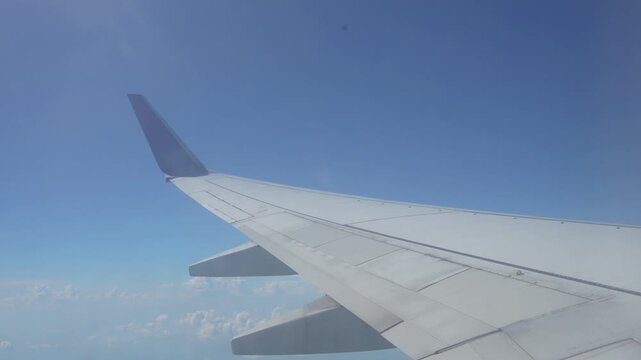 Cinematic footage of airplane wing flying in clear blue sky with soft white clouds below. Aerial view from airplane window during daytime flight.