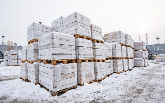 Stacked wooden pallets wrapped in white protective plastic film sit in snowy outdoor warehouse area. Large industrial cargo loads wrapped for winter protection wait for transport.