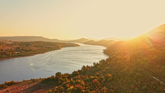 The river curves between wooded hills and fields at sunset, with the sun low on the horizon and autumn foliage covering the slopes.