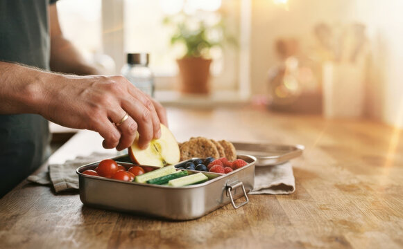Man preparing healthy lunch box with fresh vegetables, fruits, and crackers in kitchen for work or school