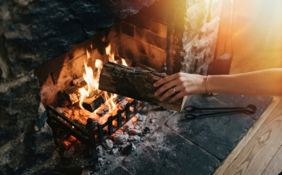 Young woman adding a log of firewood into a cozy warm fireplace indoors at home during winter evening