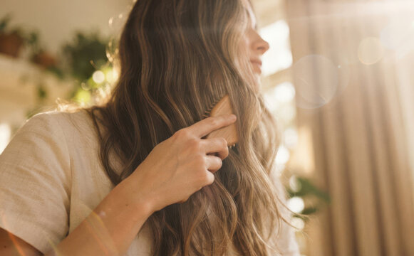 Young adult woman brushing her long wavy brown hair with wooden hairbrush at home, morning hair care routine and natural beauty concept