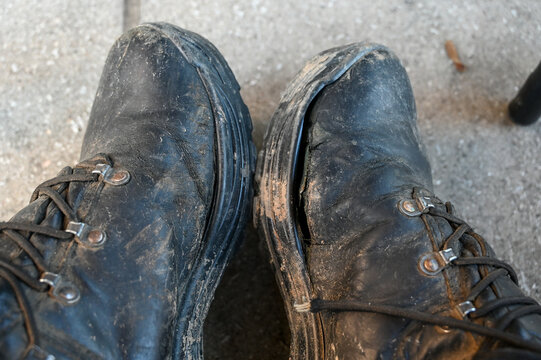 Pair of heavily worn, weathered, and caked-in-mud black leather military boots. Old, cracked and battered work boots covered in dried mud and deep creases from heavy labor. Rugged utility boots