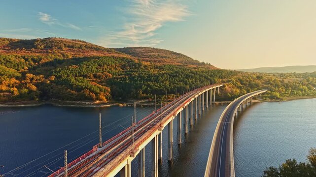 Railway tracks and a road cross a lake on separate bridges near wooded hills in autumn, with sunlight on the landscape.