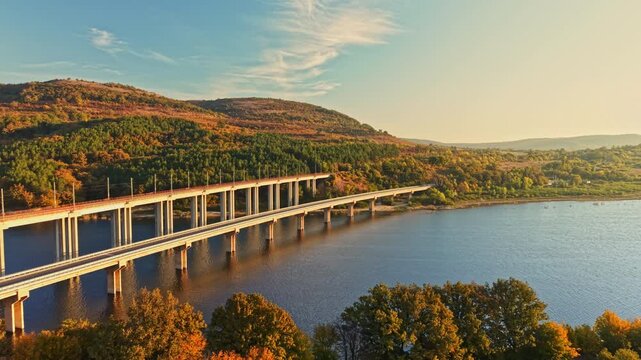 A long bridge spans a river beside wooded hills in autumn, with trees along the banks and sunlight on the landscape during the day.
