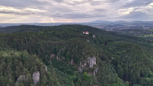 Drone aerial view over dense pine forest weaving across rolling hills to rocky outcrop with small red-roofed castle perched high above valley. Distant town and fields unfold beneath dramatic