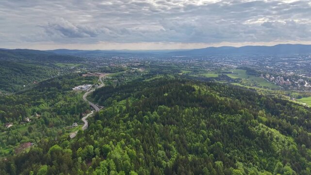 Aerial panorama shows lush rolling hills with winding road weaving through dense forest and open clearings, leading toward distant town under soft, overcast daytime sky. Wide valley, hill ranges