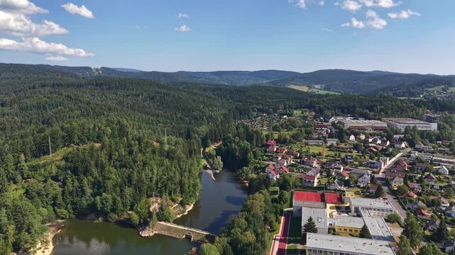 Drone captures steady aerial over tranquil river cutting through small town, surrounded by forested hills under bright sun. Sunlit rooftops, winding streets, and lush greenery create calm