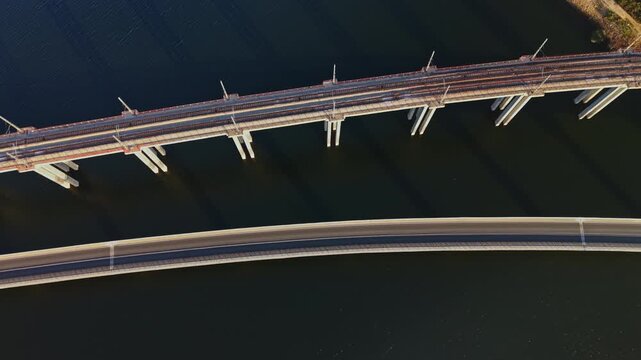 Two bridges cross a body of water, viewed from above during daylight, with vehicles moving along the upper bridge and the lower bridge spanning the frame.