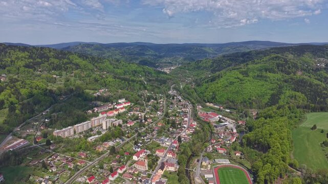 Drone glides over lush hillside town set in green valley. Wide shot captures red-roofed houses, winding streets, dense forests, and bright stadium nestled along edge of open fields. Sunlight, p