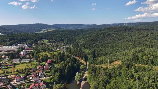 Aerial view reveals picturesque village tucked into lush green valley, framed by winding river that threads through landscape. Dense pine forests blanket rolling hills under clear blue sky