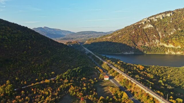 A railway line runs beside a reservoir through wooded hills near a mountain ridge, seen from above in autumn daylight.