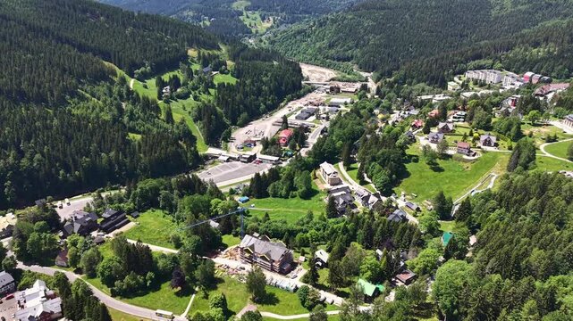 Birds-eye view of alpine village tucked in pine forested valley. Active construction site with crane near center, surrounded by winding roads, scattered houses, and lush green meadows under bright
