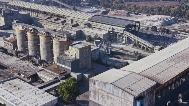 An aerial view shows an industrial plant with rows of storage silos, large factory buildings, and paved service roads during daylight.