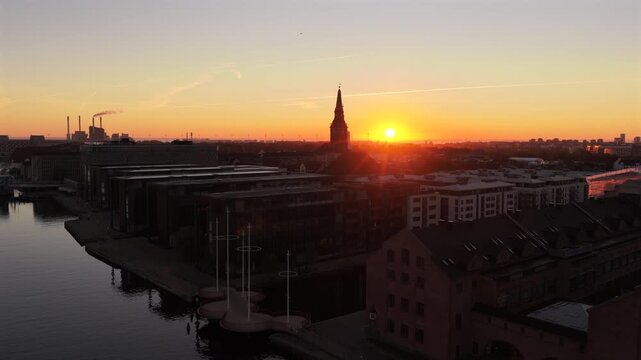Aerial drone view of the sun rising behind church spires over a harbor canal and waterfront warehouses, Copenhagen, Denmark.