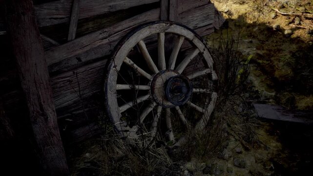 An old wooden wagon wheel leans against a decrepit barn, surrounded by tall grass and gravel. The setting evokes memories of past travels and rustic charm in a quiet countryside.