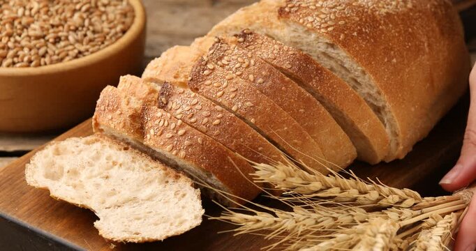 Woman putting bowl with wheat grains and spikes near fresh bread on table, closeup