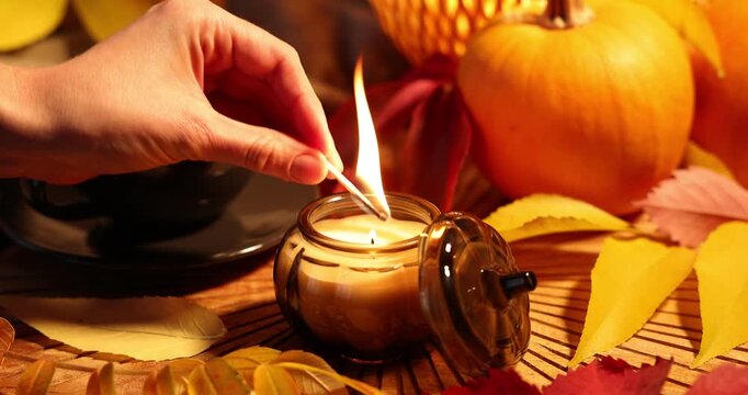 Woman lighting candle with match at table with cup of coffee, pumpkin and dry leaves, closeup. Cozy autumn atmosphere