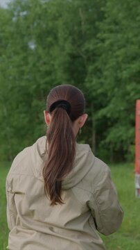 Woman stands outdoors her back to camera wearing beige jacket and ponytail in grass field with trees. orange object is visible on right side of some frames