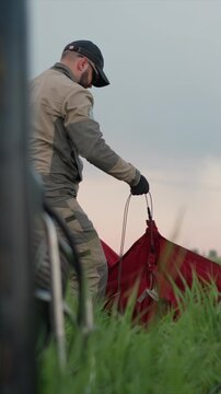 Woman and man in beige jumpsuits prepare parachuting equipment on grassy field under cloudy sky. man handles ropes attached to red parachute bag while woman assists with setup
