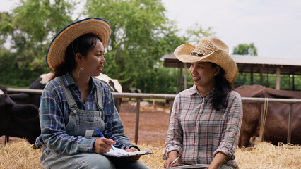 Two female dairy farmers sit beside hay bales, discussing cattle farm operations while one writes notes on a clipboard and the other holds a tablet, highlighting teamwork and livestock recordkeeping. © Father_Studio