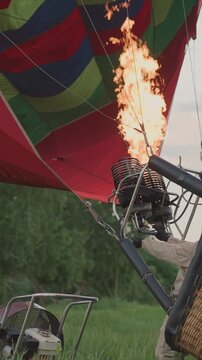 Hot air balloon burner flame ignites as pilot in tan uniform operates equipment with wickered basket and surrounding green field visible. scene captures preparation details for aerial ascent