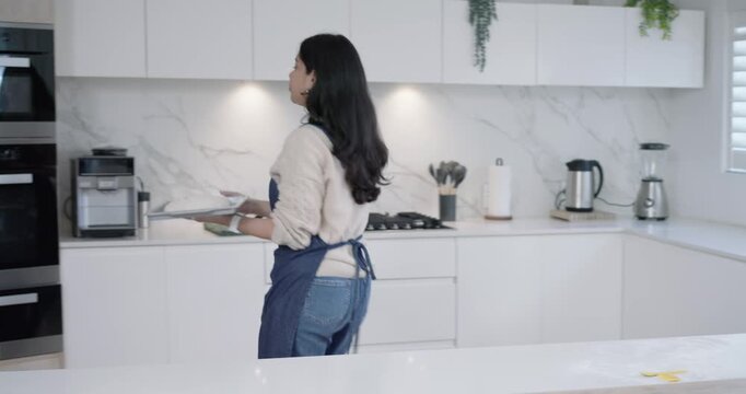 Woman shaping dough with small yellow tool, carrying tray across countertop into oven for baking