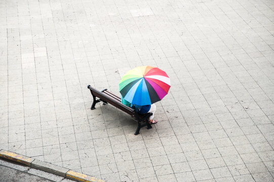 minimalist image viewed from above of person sat on a square bench with rainbow umbrella viewed from above