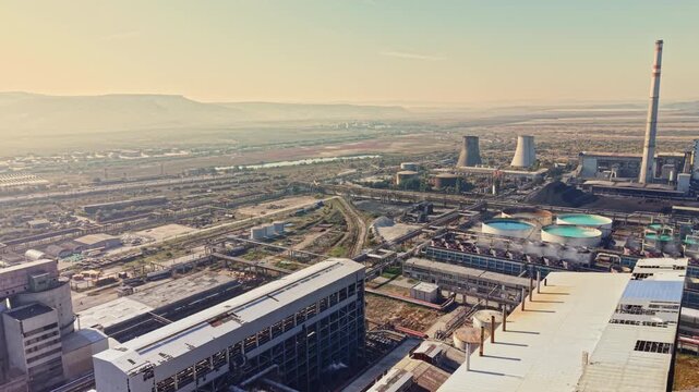 Power plant structures, smokestacks, cooling towers, and factory buildings extend across an industrial site beneath a hazy sky during the day.