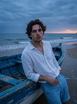 A Young Man in a White Linen Shirt Leaning Against a Blue Boat on the Beach at Dusk